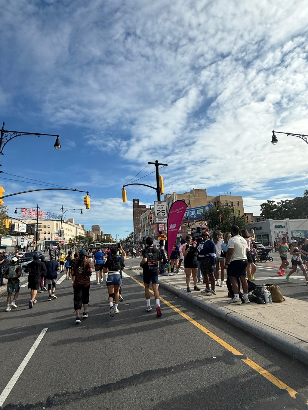 Back-of-the-pack runners heading up the Grand Concourse in the Bronx under a wide blue sky, with spectators cheering along the curb