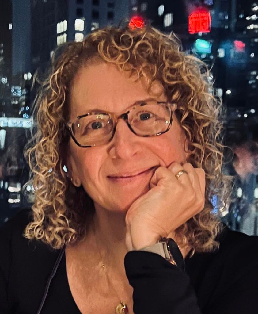 Iris Derke smiling, chin resting on her hand, with the lights of New York City at night behind her