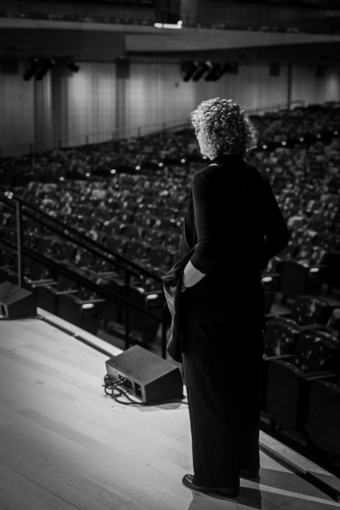 Black and white photo of Iris Derke standing alone at the edge of the stage at David Geffen Hall, looking out over the empty seats