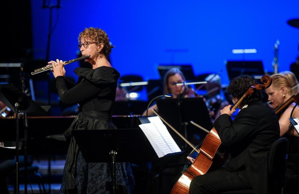 Iris Derke playing flute on stage with an orchestra under deep blue light